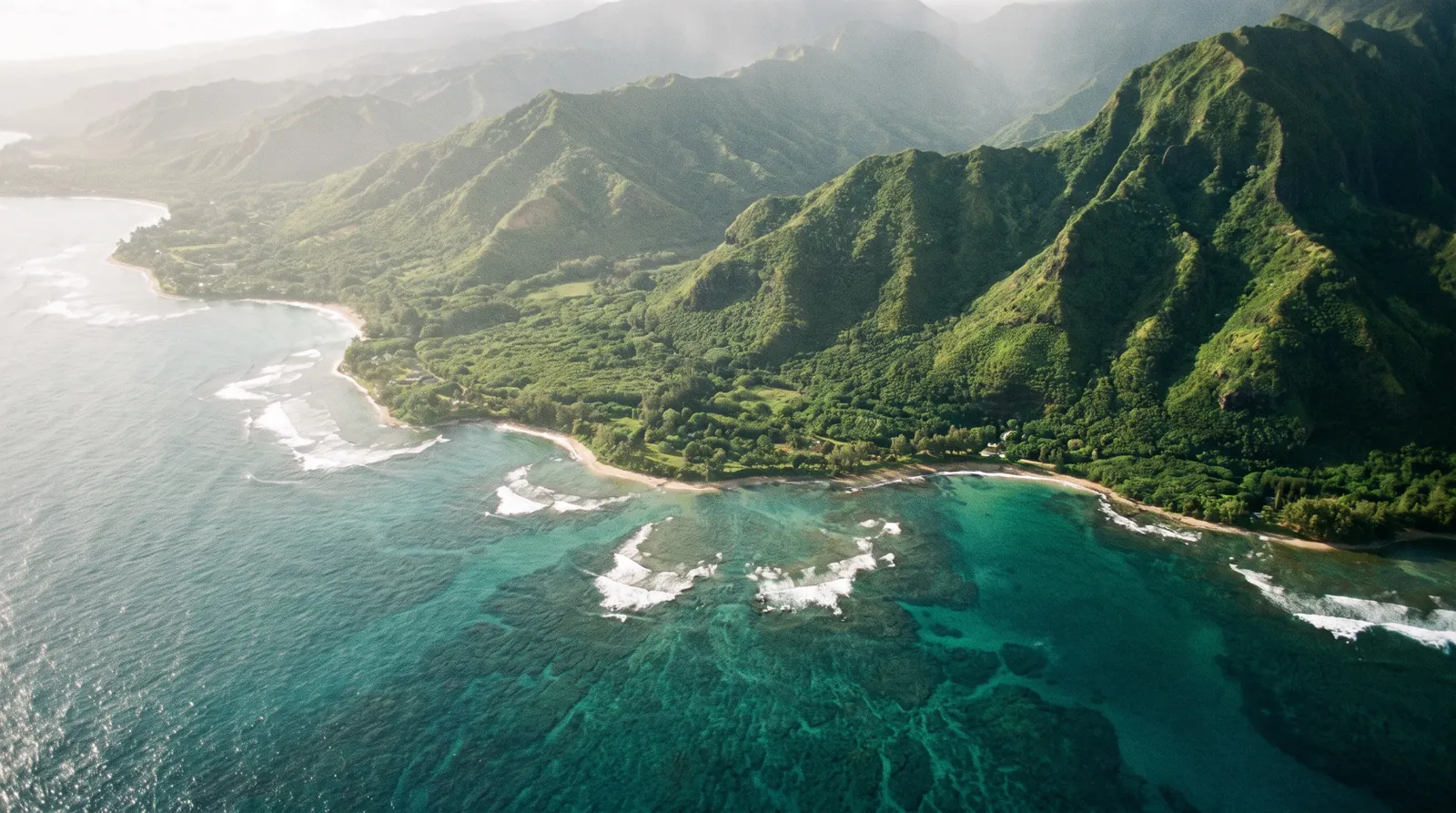 Aerial view over a Maui coastline — reef-streaked turquoise water meeting deep blue, green ridges rising inland